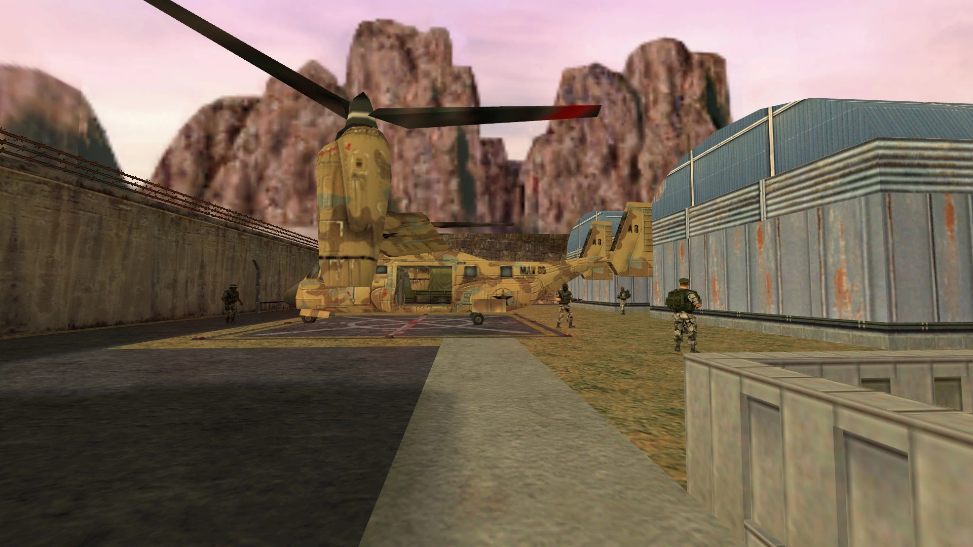 Getarnter Militärhubschrauber auf einer Landefläche mit Soldaten neben einer großen Industriehalle und felsigem Hintergrund