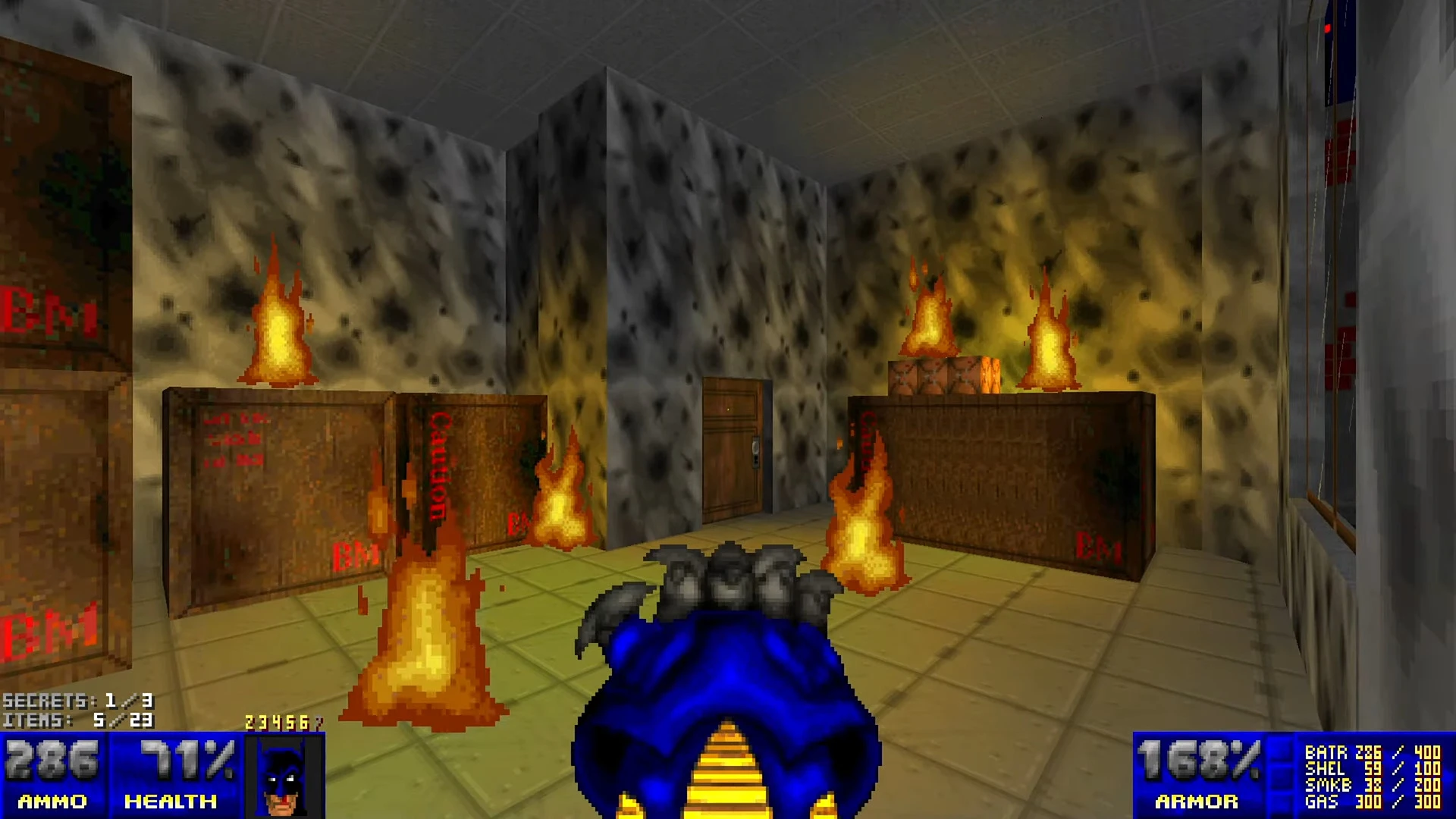 First-person view of a blue weapon inside a smoky room with wooden crates on fire in Doom II Batman mod