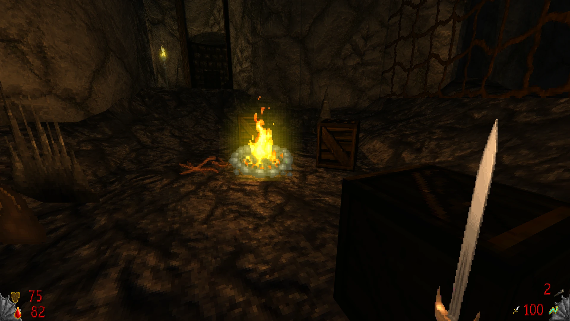 Ігровий вигляд печери з кам'яними стінами, багаттям у центрі, дерев'яними ящиками та мечем у руці