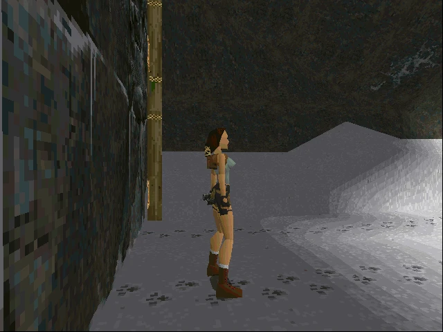 Lara Croft de pie dentro de una cueva tenuemente iluminada con paredes y suelo rocosos en Tomb Raider 1996