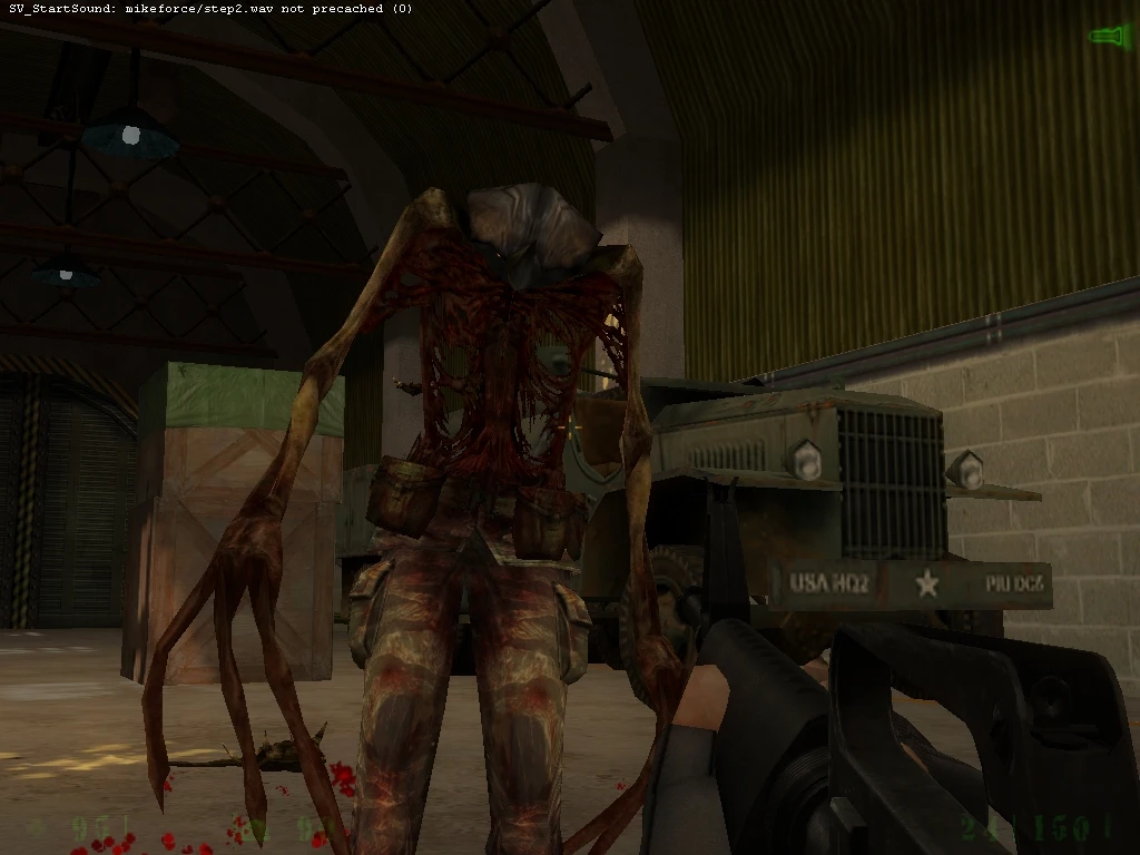 In-game screenshot showing a first-person shooter view aiming at a grotesque alien creature inside a military hangar with crates and a jeep